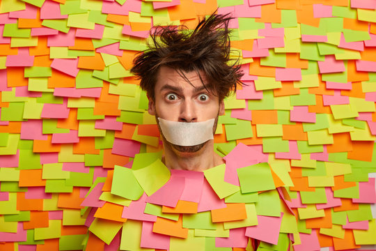 Surprised nervous man with adhesive duct tape over mouth, asks be silent, stands silenced and speechless, poses against colorful background with sticky notes, being frightened. Shut up, censorship