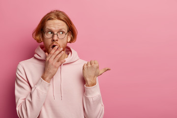 Wall Mural - Shocked concerned ginger hipster covers opened mouth, expresses amazement, shows something disturbing, stands astounded, points thumb aside, tells you urgent shocking news, wears sweatshirt.