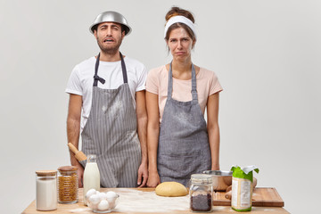 Wall Mural - Tired discontent housewife and husband stand together at kitchen near table with ingredients, make dinner for family, prepare dough for baking pizza, exhausted of routine activity, wear aprons