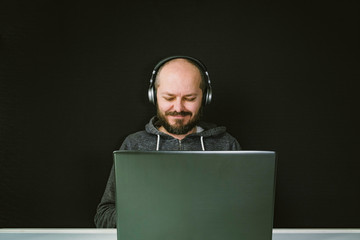 Caucasian man in hoodie and headphones sitting behind the laptop and working, smiling, black background, home office concept