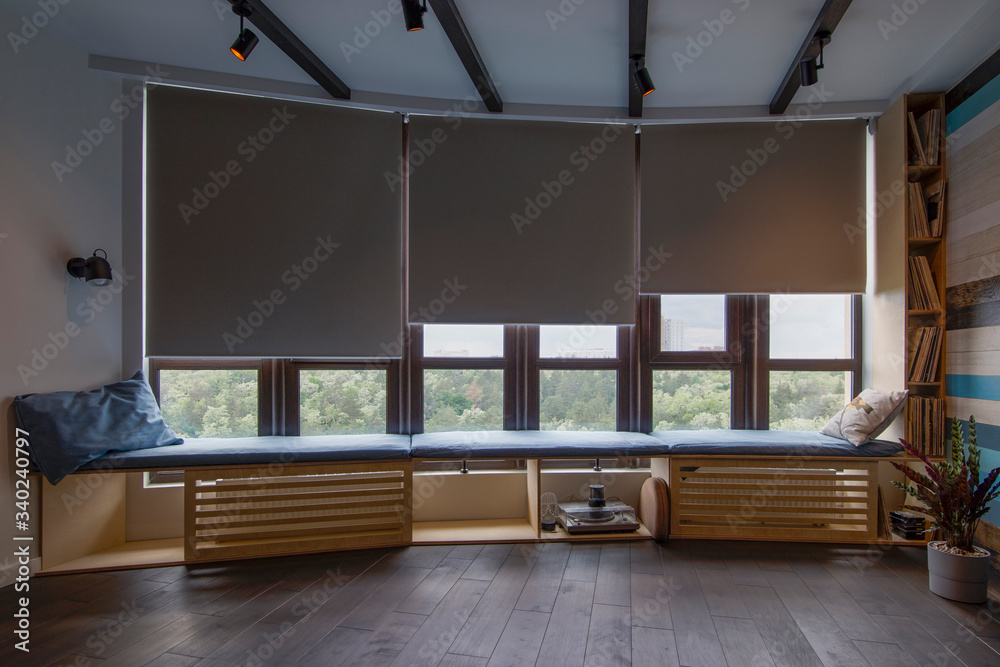 Motorized roller shades in the interior. Automatic roller blinds beige color on big glass windows. Remote Control Shades are above the windosill with pillows. Summer. Green trees outside. - obrazy, fototapety, plakaty 