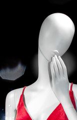 Eyeless, noseless, hairless white mannequin in red dress with hand over mouth in black background