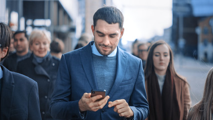 Wall Mural - Caucasian Businessman in a Suit is Using a Smartphone on a Street in Downtown. Other Office People Commute in a Crowd. He's Confident and Looks Successful. He's Browsing the Web on his Device.