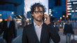 © Gorodenkoff - Young Stylish Businessman with Curly Hair is Talking on a Phone While Walking in the City. He's Wearing Glasses. Office People Walk By. It's Evening with Atmospheric Urban Lights in the Background.