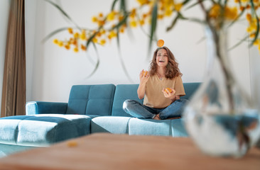 Charming beautiful young girl with curly hair and a happy smile juggles tangerines while sitting on a blue sofa in her living room at home, having fun staying at home