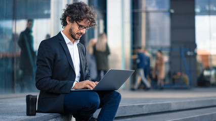 Wall Mural - Handsome Businessman in a Suit is Sitting on Steps next to Business Center and Working on a Laptop on a Street in a City. Office People Walk By to Work.