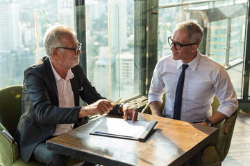 Picture of two business man discussing business on a table.  The young man is wearing a white shire with black tie and glass. The older man wear a black suit and sunglass. They are in a hotel lobby.