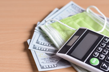 Close-up  of one hundred dollars   and  medical disposable  face mask   with   handheld mini calculator  on wooden background . The economic impact of coronavirus  in  United States of America .