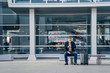 © VK Studio - Outdoor shot of male passenger waits for transport on bench, poses at station, looks at watch, suitcase on wheels near, wears medical mask during quarantine and coronavirus spread. Traveling