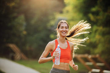 Wall Mural - Young smiling positive attractive sportswoman with long hair holding bottle of water and running in public park in the morning.