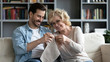 © fizkes - Grown up son visit elderly mother, different generation family sit on couch, millennial guy adult child try to knit use needles and yarn aged mom teaches assist him, people spend funny time together