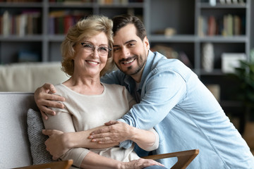 Younger and older generation relatives people portrait, love, care, connection, family bonds concept. Grown up adult son hugs elderly mother while she resting seated on cozy comfort armchair indoors