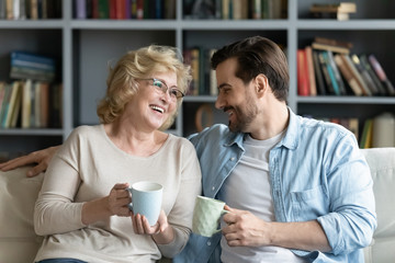 Old mom and grown up son sitting on sofa holding cups with hot beverage drinking tea enjoy warm friendly conversation, sharing news, remember pleasant moments of life, connection familial ties concept