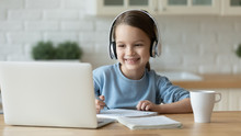 Smiling Little Caucasian Girl In Headphones Handwrite Study Online Using Laptop At Home, Cute Happy Small Child In Earphones Take Internet Web Lesson Or Class On Computer, Homeschooling Concept