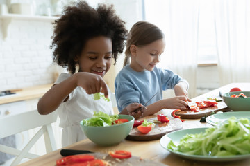 Wall Mural - Excited little multiracial girls sisters have fun chopping vegetables making healthy salad at home, happy small multiethnic siblings children cooking in kitchen together, preparing food for parents