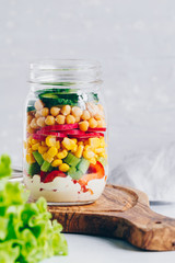 Wall Mural - Layering salad in a glass jar. Fresh radish, cucumber, celery, red bell pepper, corn, chickpeas and sauce.