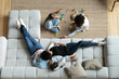 © fizkes - Top view of happy multiethnic young family with small children relax in living room at home together, diverse parents rest on comfortable sofa watch multiracial little daughters play on warm floor