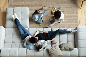 Top view of happy multiethnic young family with small children relax in living room at home together, diverse parents rest on comfortable sofa watch multiracial little daughters play on warm floor