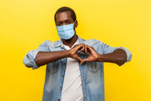 Romantic Relations. Portrait Of Handsome Cheerful Man With Surgical Medical Mask Making Heart Shape With Hands, Expressing Love Feelings Or Friendship. Indoor Studio Shot Isolated On Yellow Background
