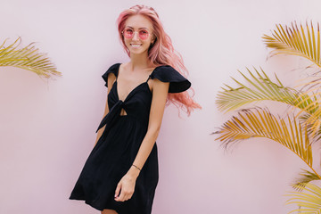 Wall Mural - Lovely tanned girl in black dress posing in studio and laughing. Indoor portrait of carefree caucasian woman with pink hair standing near palm trees.