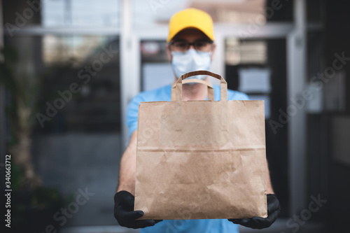 Delivery guy with protective mask and gloves holding box / bag with groceries in front of a building.