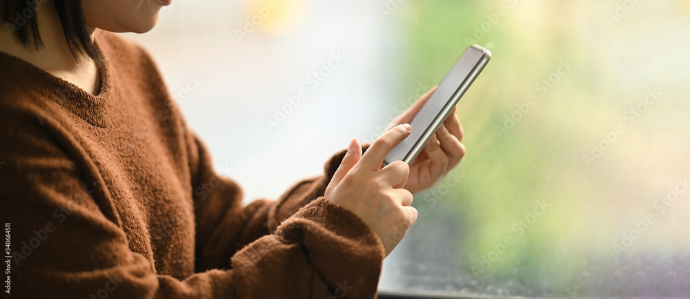 Cropped image of beautiful woman on brown cotton sweater holding a white blank screen smartphone in her hands while sitting and relaxing on her holidays in comfortable sitting room as background. - obrazy, fototapety, plakaty 