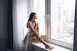 © Алина Троева - Beautiful bored slender brunette woman sitting next to window windowsill under warm grey blanket at home. Self isolation quarantine during Corona virus pandemic. COVID19 stay home save lives concept