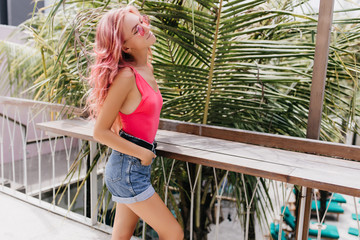 Wall Mural - Shapely glamorous woman posing with eyes closed in front of palm tree. Outdoor photo of pensive girl with pink hair wears tank-top and denim shorts.