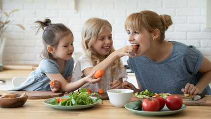 caring daughter feeding mother teaching little children standing at table in kitchen. happy mom with