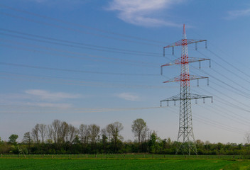 high voltage power lines, red and white electricity pylon with blue sky and green field with trees, symbol for infrastructure, energy and nature