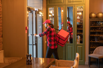 Wall Mural - Young busy courier with big bag opening the door while going to deliver orders