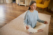 © shurkin_son - High angle view of gray haired mature female in casual clothes sitting on carpet in half letter pose, keeping eyes closed, practicing meditation, letting go thoughts, concentrating on breathing