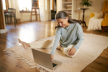 Indoor image of beautiful energetic female on retirement sitting barefoot on floor using laptop turning on calm music for meditation. Elderly European woman surfing internet on portable computer