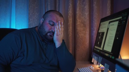 Overworked tired young fat man freelancer sitting at computer in night, home office. Online distant work concept in modern world