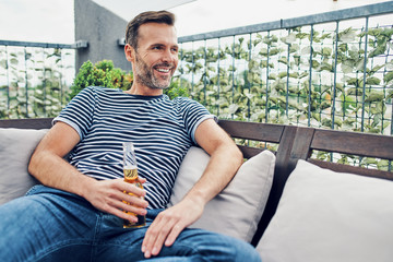 Portrait of cheerful man sitting on patio with bottle of beer and relaxing