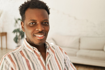 Wall Mural - Close up image of cool cheerful young dark skinned man with Afro hairstyle sitting in stylish living room interior, relaxing at home, looking at camera with broad happy smile. People and lifestyle