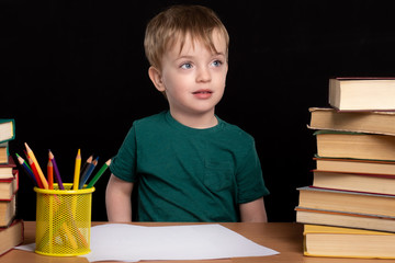 little boy sits at a desk with books and pencils. Black background. isolate. look to the side. close-up portrait.