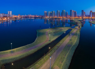 Wall Mural - Night view of the city from the drone. A deserted walking area with lakes on the background of a residential complex. Apartments in new buildings. Developed urban infrastructure.
