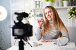 © artiemedvedev - Vlogger female showing lipstick. Beauty blogger woman filming daily make-up routine tutorial near camera on tripod. Influencer blonde girl live streaming cosmetics product review in home studio