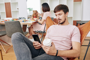 Wall Mural - Handsome young man drinking coffee and texting friends when his wife is playing with daughter