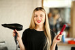Young Hairstylist Holding Blow Dryer and Hairbrush. Blonde Hairdresser Showing Hairdryer and Red Comb for Haircut Styling. Stylist Using Professional Equipment for Making Hairdo Looking at Camera Shot