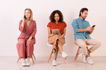 Wall Mural - Modern multi-ethnic people in casual outfits sitting on chairs in row and using gadgets all time for communication