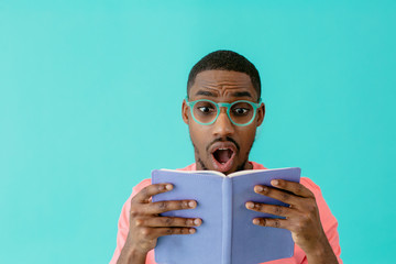 Portrait of a young person shocked at what he is reading holding book, isolated on blue copy space