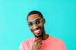 © Carlos David - Portrait of a happy young man with glasses smiling, thinking and looking at camera with hand on chin, against blue studio background