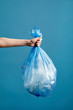 © Seventyfour - Vertical image of female hand holding trash bag with plastic against blue background, waste sorting and recycling concept, copy space