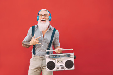 Senior crazy man listening music with headphones and vintage boombox outdoor - Hipster male having fun living in past time - Elderly people lifestyle activity - Red background