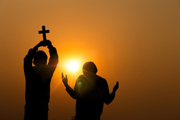Wall Mural - Two young christian holding Cross and lift hands up to sky praying to God with light sunset background, Christians should worship and thank God, christian silhouette concept.
