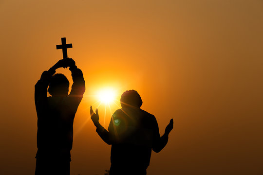 Two young christian holding Cross and lift hands up to sky praying to God with light sunset background, Christians should worship and thank God, christian silhouette concept.