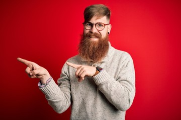 Canvas Print - Handsome Irish redhead man with beard wearing casual sweater and glasses over red background smiling and looking at the camera pointing with two hands and fingers to the side.