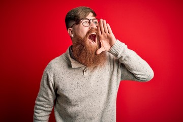 Poster - Handsome Irish redhead man with beard wearing casual sweater and glasses over red background shouting and screaming loud to side with hand on mouth. Communication concept.